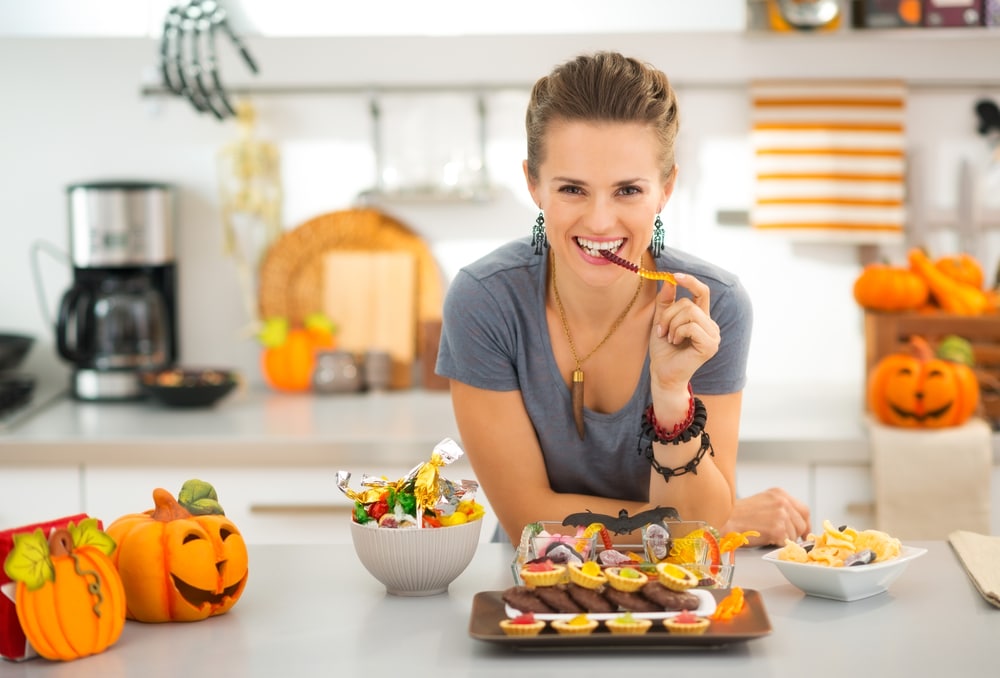 Ready to halloween party! Smiling woman in decorated kitchen eating halloween trick or treat candy